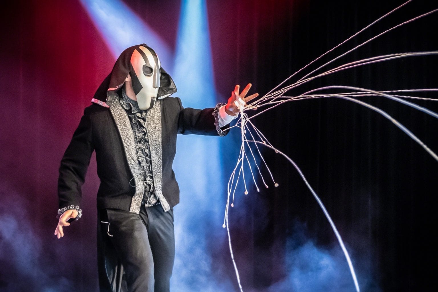 Daniel Kane i mask och glittrande kavaj med lysande trådar som sprakar ut ur handen i blått och rött scenljus